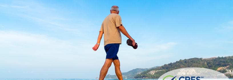 senior male holding flip flops walking along ocean edge with tree covered hills in the background