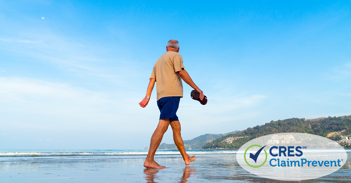 senior male holding flip flops walking along ocean edge with tree covered hills in the background